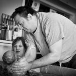 Mann hält Neugeborenes über Badewanne neben Mutter